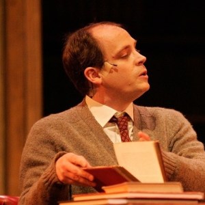 A man in a brown cardigan opening a large book.
