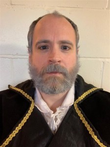 A man wearing a brocaded jacket. He has a bushy grey beard.